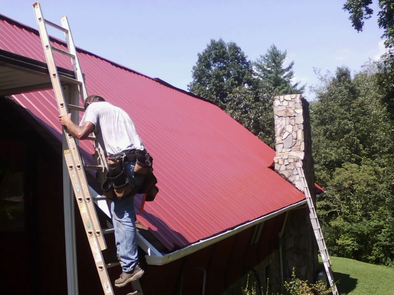 Roofer on a ladder beside a red metal roof and stone chimney — Soffit & Fascia Repair by Roofing Brothers in Turney